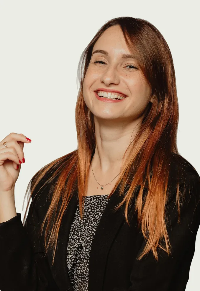 Smiling woman with long reddish hair wearing a black blazer and patterned top against a plain background.