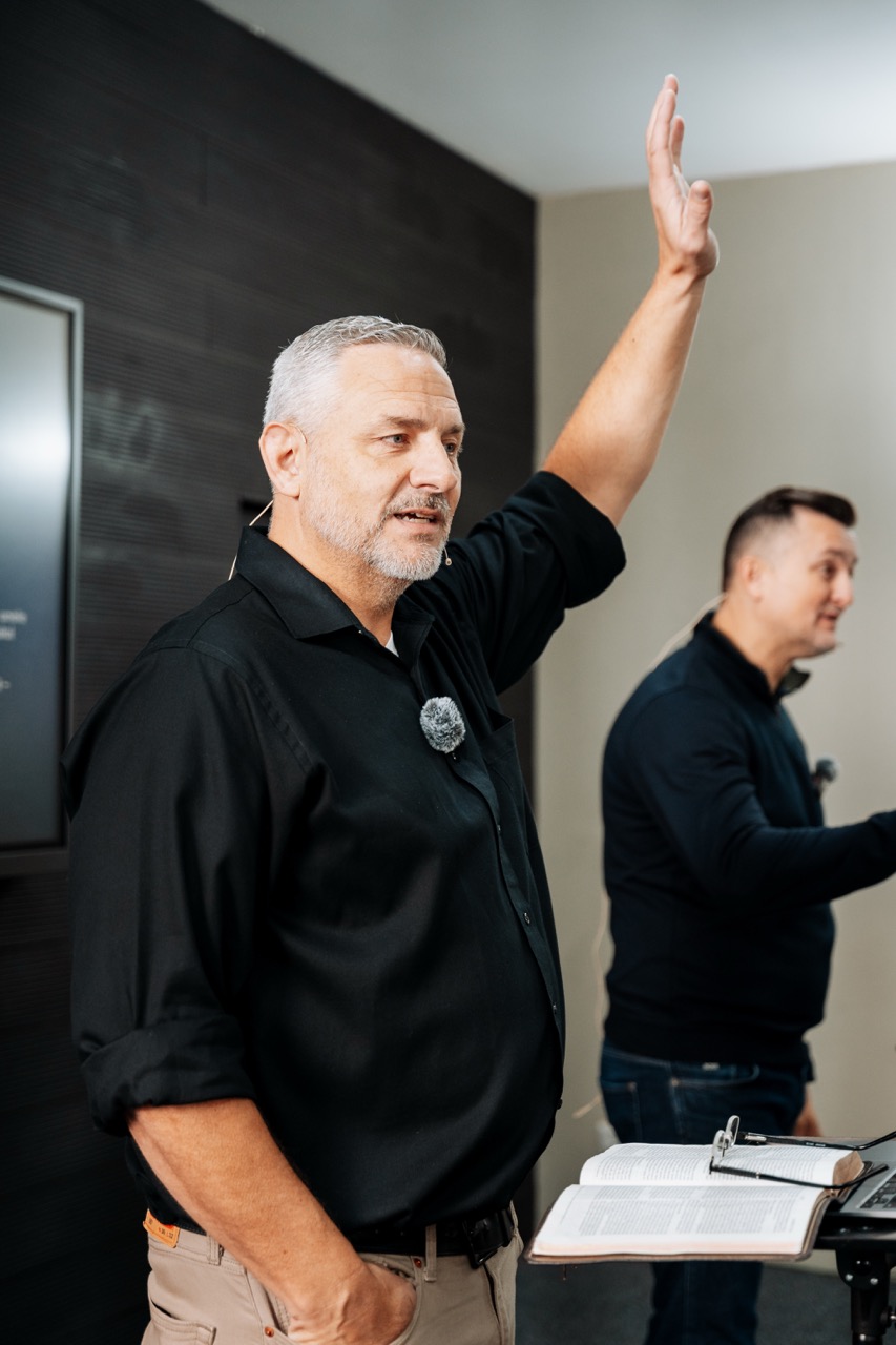 Man with gray hair and beard raising his hand while speaking in front of a lectern with an open book, another man in the background also speaking.