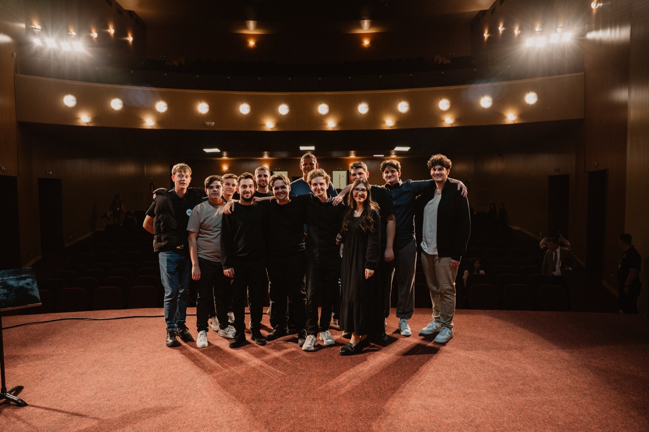 Group of twelve young adults standing closely together on a theater stage under warm lights.
