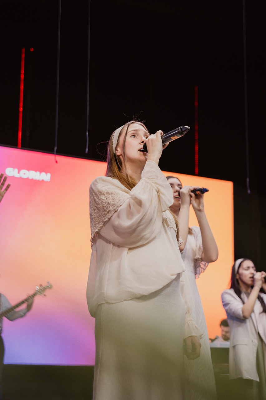 Women singing on stage with microphones in front of colorful illuminated screen displaying the word 'GLORIA!'