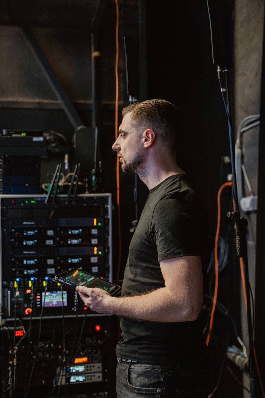 Man in black t-shirt holding a tablet, standing next to audio and networking equipment with cables in a dimly lit room.