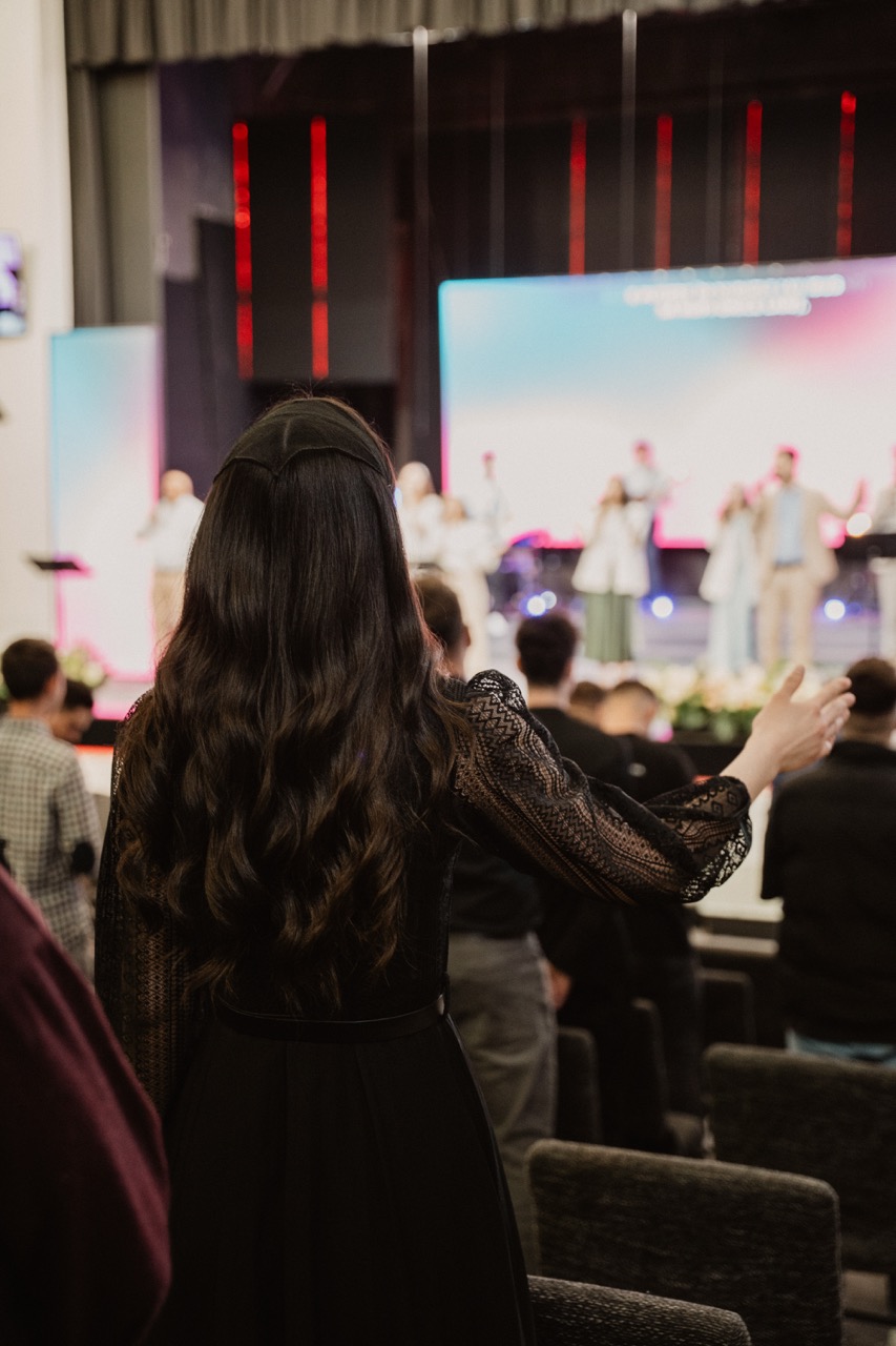 Woman with long dark hair and a black lace dress raising her hand in a crowded auditorium during a stage performance.