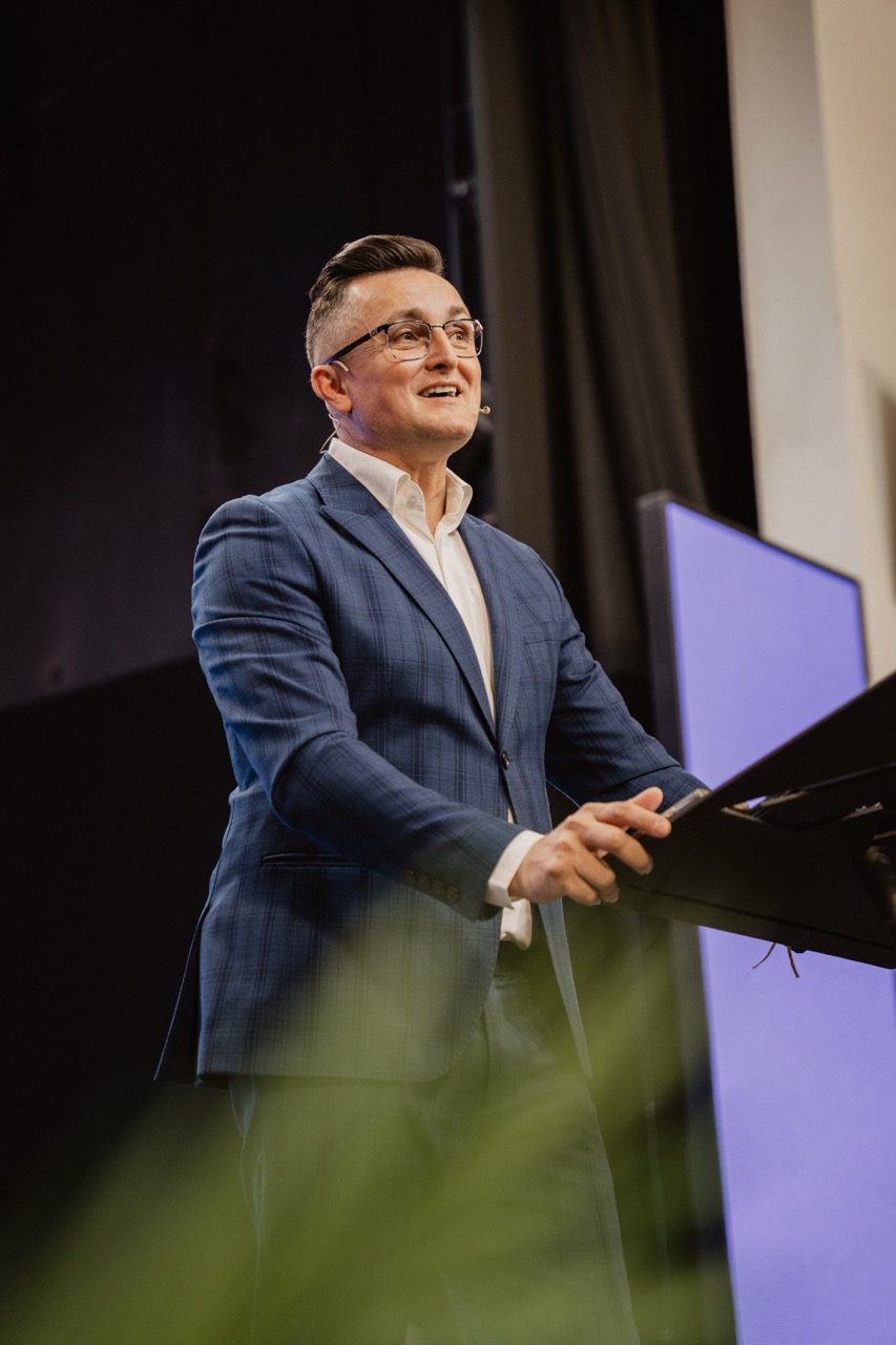 Man in a blue suit and glasses speaking at a podium with a microphone headset.