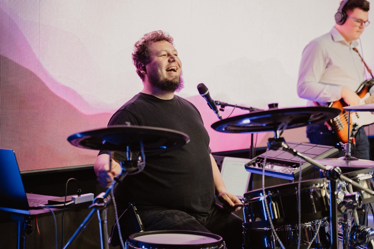 Man playing electronic drums and smiling while another man with headphones plays bass guitar in the background.