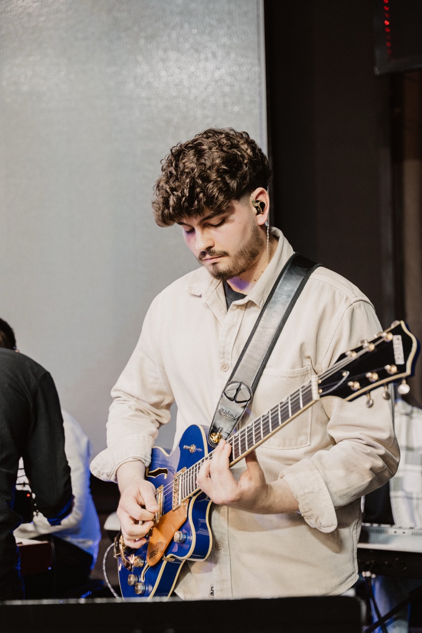 Man with curly hair playing a blue electric guitar while wearing a beige shirt and black ear monitors on stage.