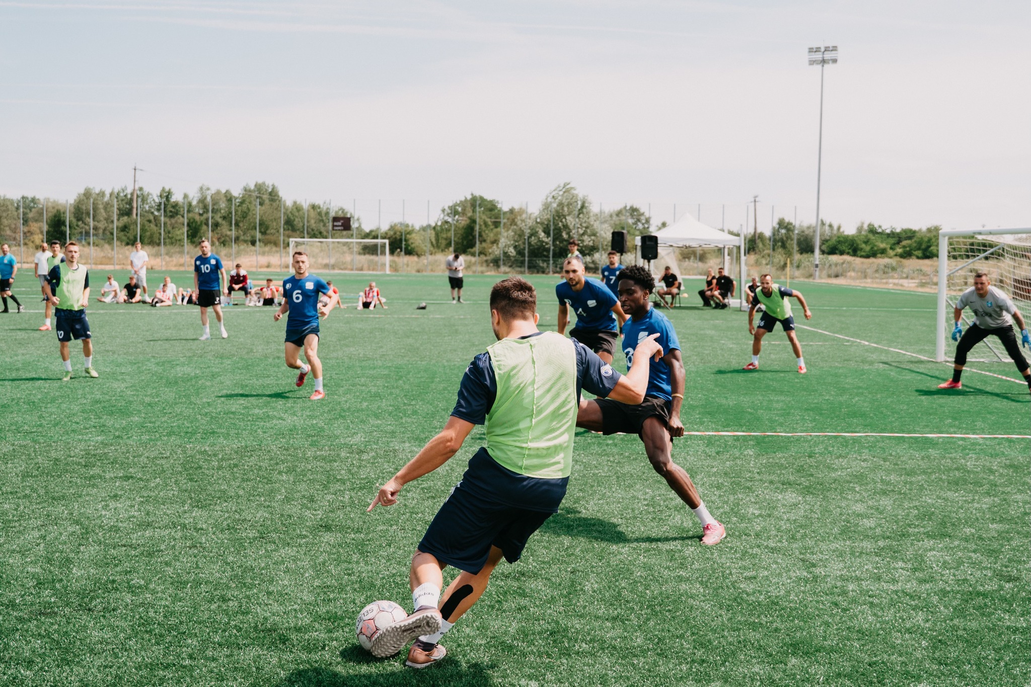 Soccer player in green vest kicking a ball while opposing player in blue defends on a sunny outdoor field.