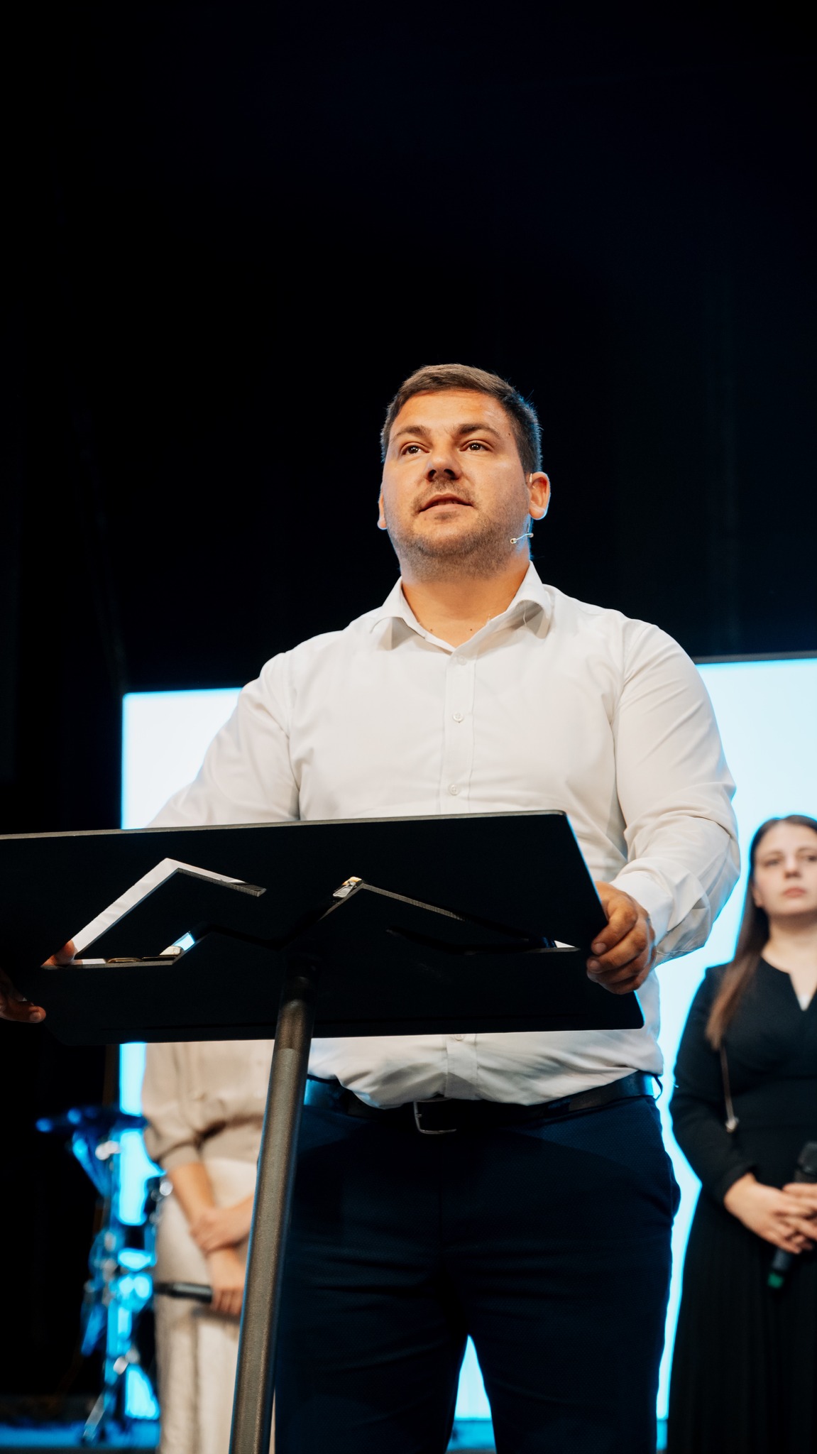 Man in white shirt speaking at a podium with two women standing behind him holding microphones.