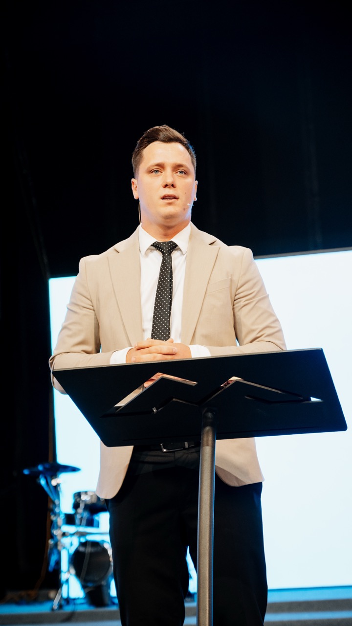 Man in beige suit jacket and dotted tie speaking behind a black podium on stage with drum set in the background.