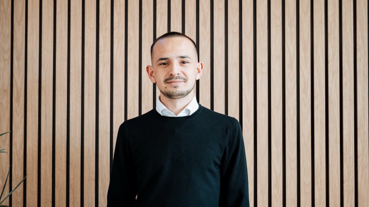 Smiling man with short hair and beard wearing a black sweater and white shirt standing in front of a wooden slat wall.