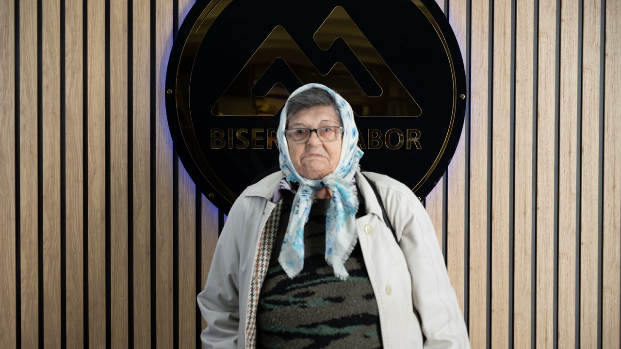 Elderly woman wearing glasses, a light jacket, and a floral headscarf standing in front of a wooden slat wall with a black circular sign.