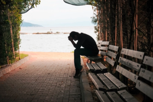 Silhouette of a person sitting alone on a bench with their head in their hands, overlooking the water, representing isolation, grief, or sadness.