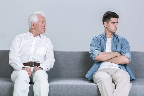 Older father looking with concern at his adult son, who sits turned away with arms crossed, representing a family disagreement or communication breakdown.
