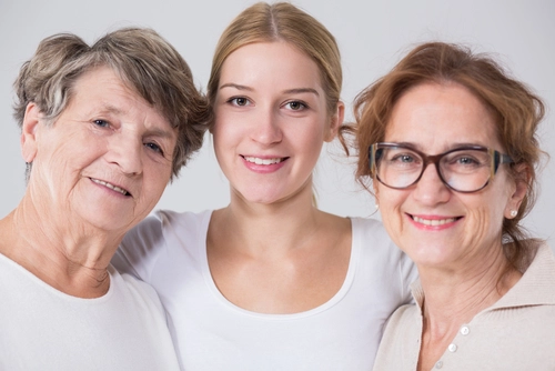 Portrait of three smiling women of different generations standing close together against a plain background.