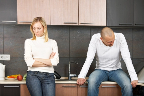 Upset couple in a kitchen; a woman stands with arms crossed looking away while a man sits on the counter looking down.