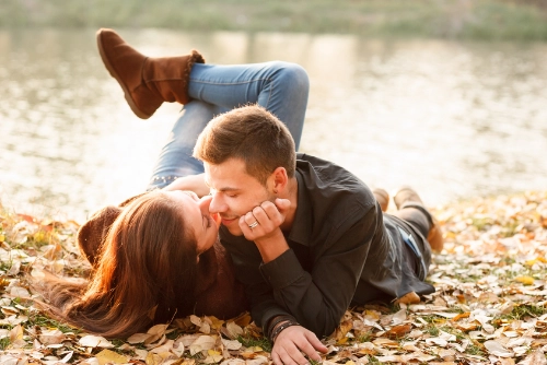 A couple lying on autumn leaves looking affectionately at each other with noses touching, representing intimacy and connection.