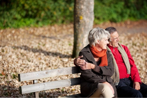 A smiling older couple sitting on a park bench together; the man has his arm around the woman, depicting long-term companionship.
