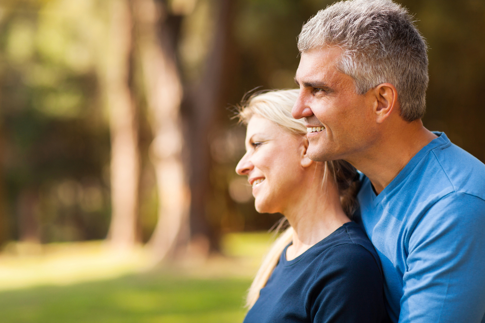 A smiling older couple sitting on a park bench together; the man has his arm around the woman, depicting long-term companionship.