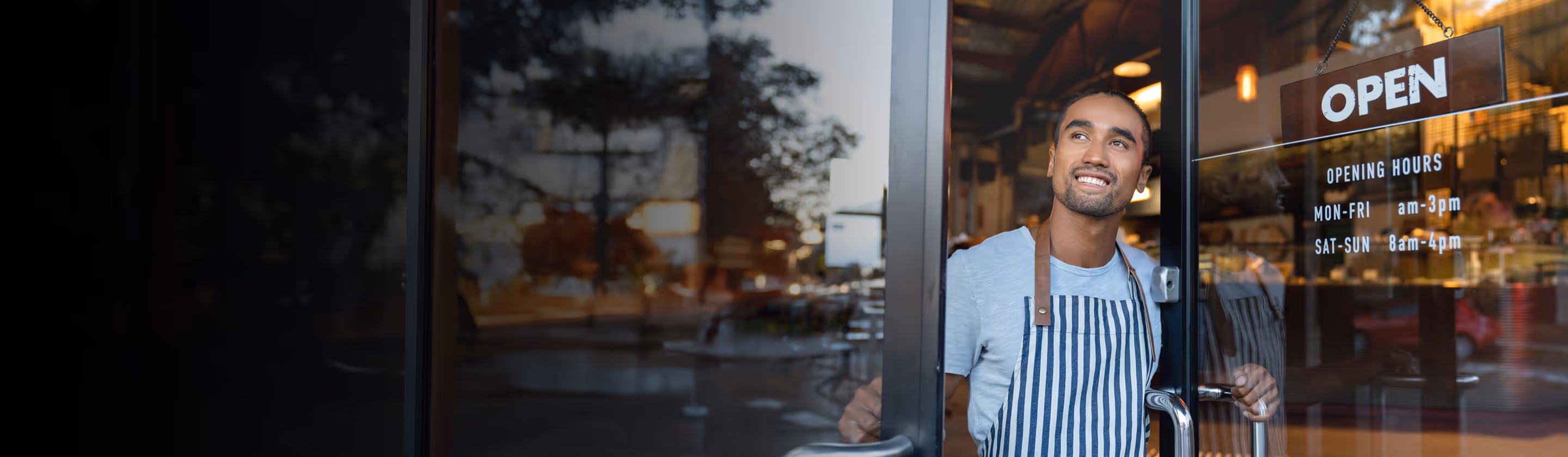 Smiling man in striped apron opening a cafe door with an open sign and business hours displayed.