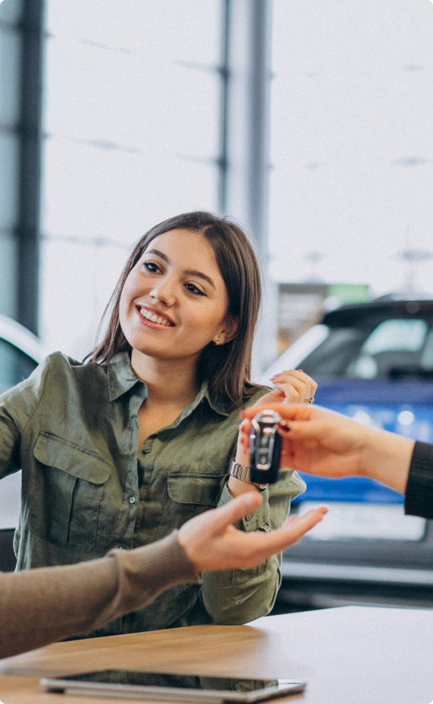 Smiling woman sitting at a dealership table receiving car keys from a salesperson after financing approval.