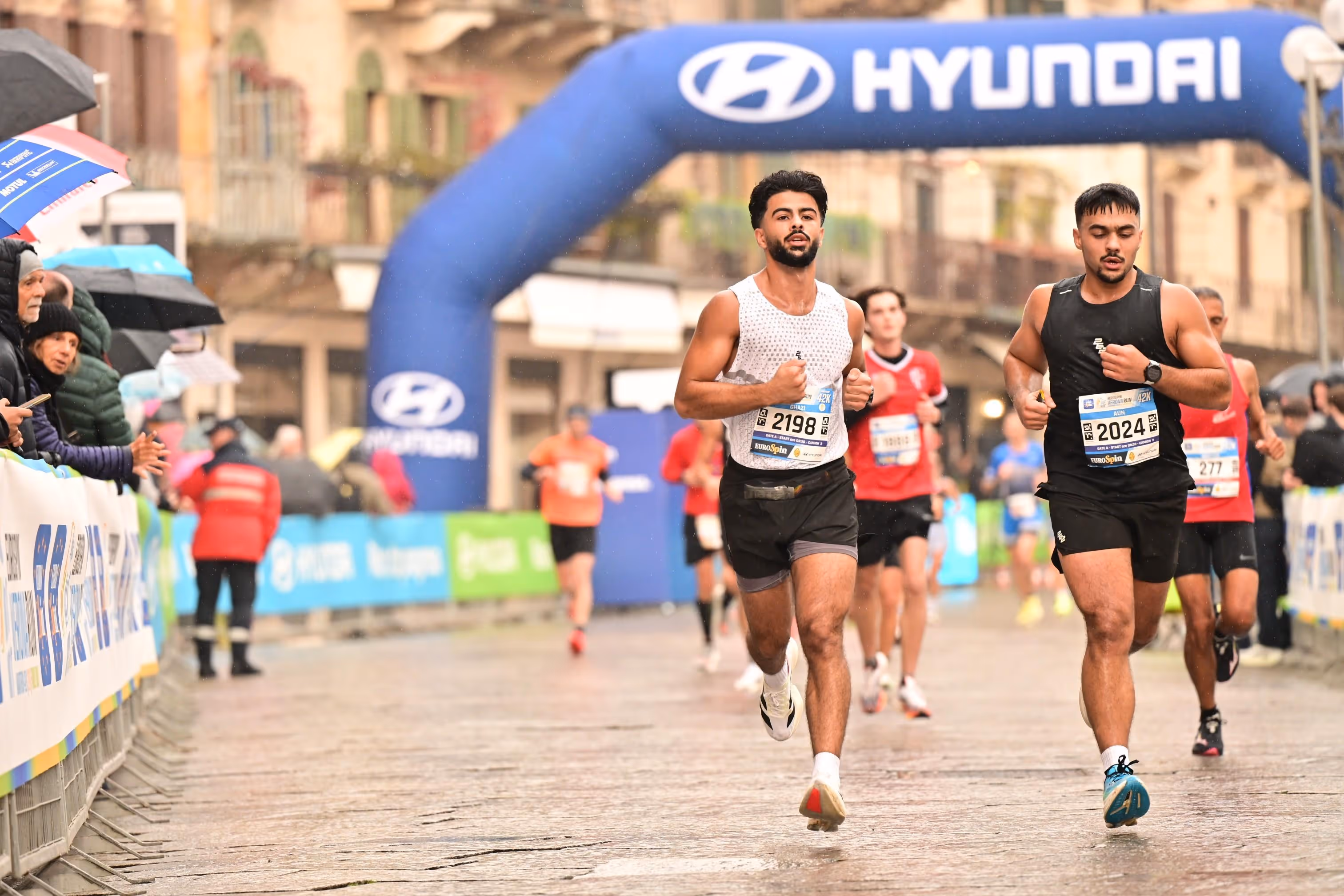 Runners competing in a race on a wet street under a blue Hyundai inflatable arch with spectators watching and holding umbrellas.