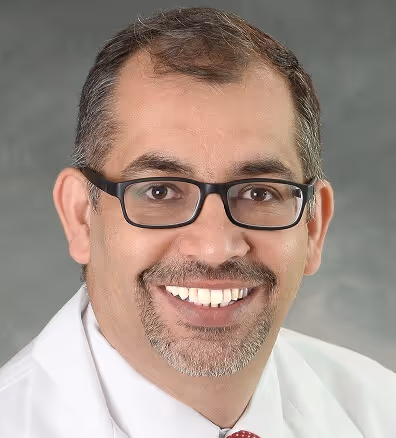 Middle-aged man with glasses, a trimmed beard, and mustache smiling, wearing a white coat and red tie against a gray background.