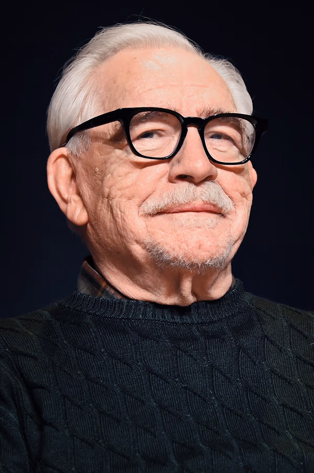 Elderly man with white hair and glasses smiling warmly in black sweater
