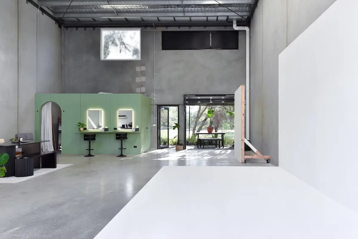 Spacious modern studio with polished concrete floors, a green makeup station with illuminated mirrors, and large open windows showing greenery outside.