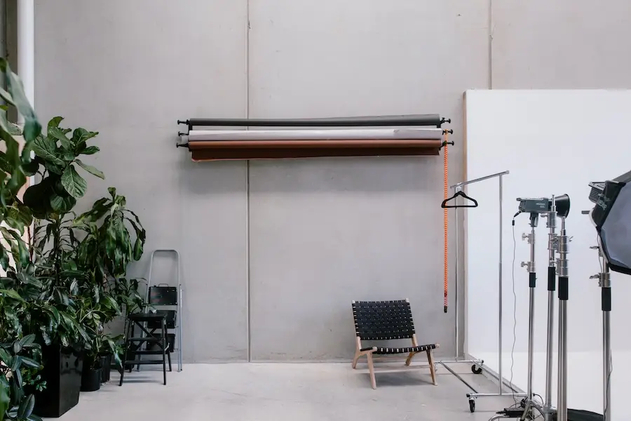 Minimalist photography studio with neutral backdrops on the wall, black woven chair, lighting equipment, clothing rack with hanger, and green plants.