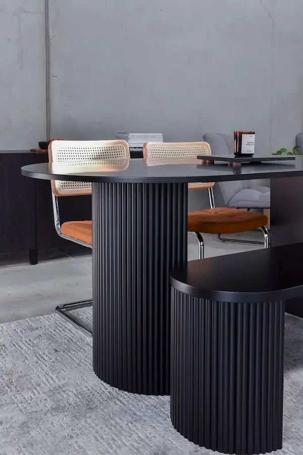 Modern black round table with vertical grooves paired with two rattan-backed chairs with brown cushions on a gray rug.