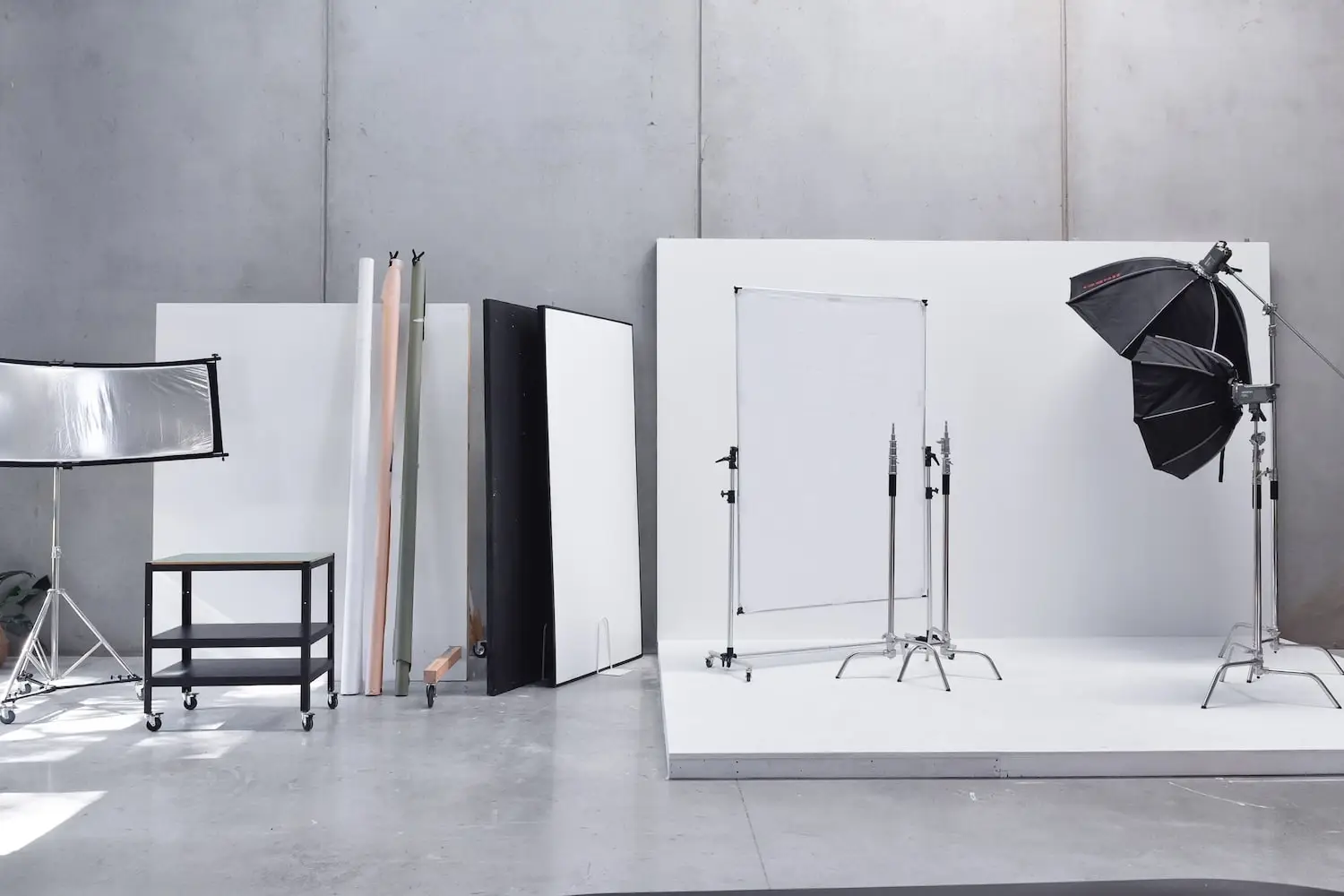 Minimalist photography studio setup with softbox lights, reflectors, and a black rolling cart against a gray concrete wall.