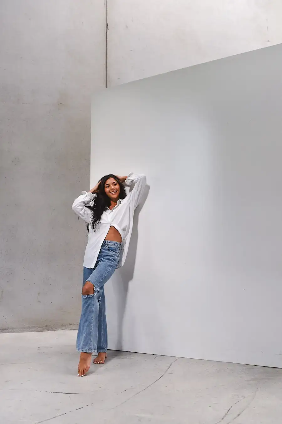 Smiling barefoot woman with long dark hair wearing a white shirt and ripped blue jeans leaning against a white wall.
