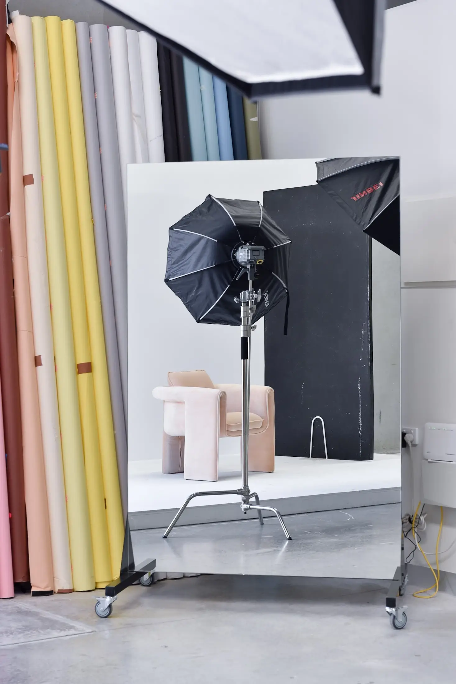 Photography studio setup with a softbox light, a black reflector, and a light pink armchair reflected in a large mirror, with various colored backdrops on the side.