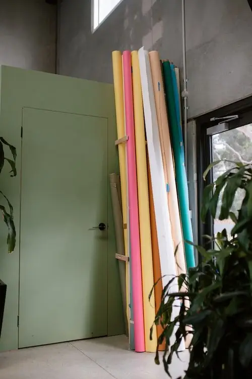 Stacks of colorful rolled paper leaning against a green door in a concrete room with plants nearby.