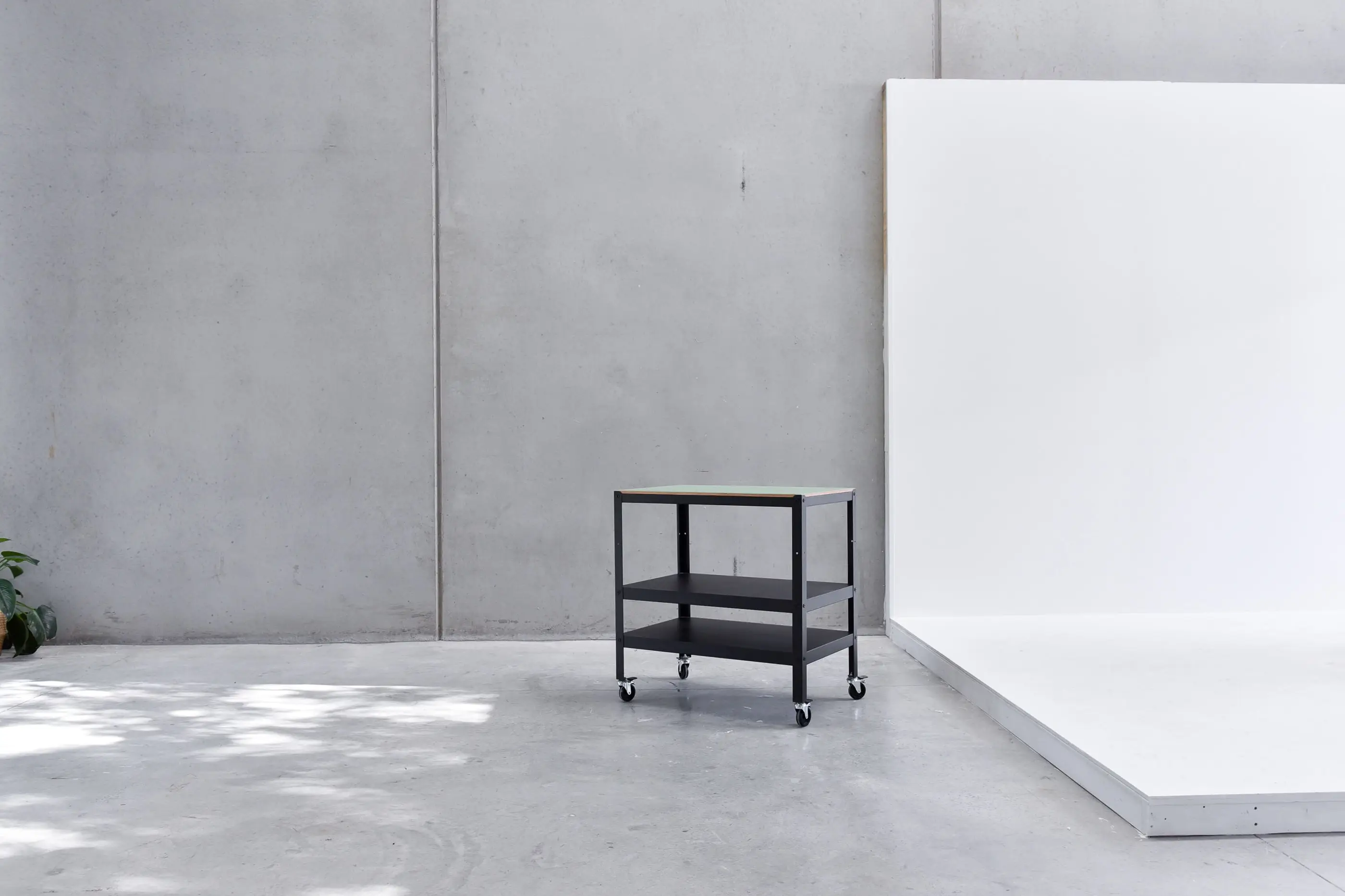 Black metal rolling cart with two shelves and a light-colored top on a concrete floor against a gray concrete wall and white platform.