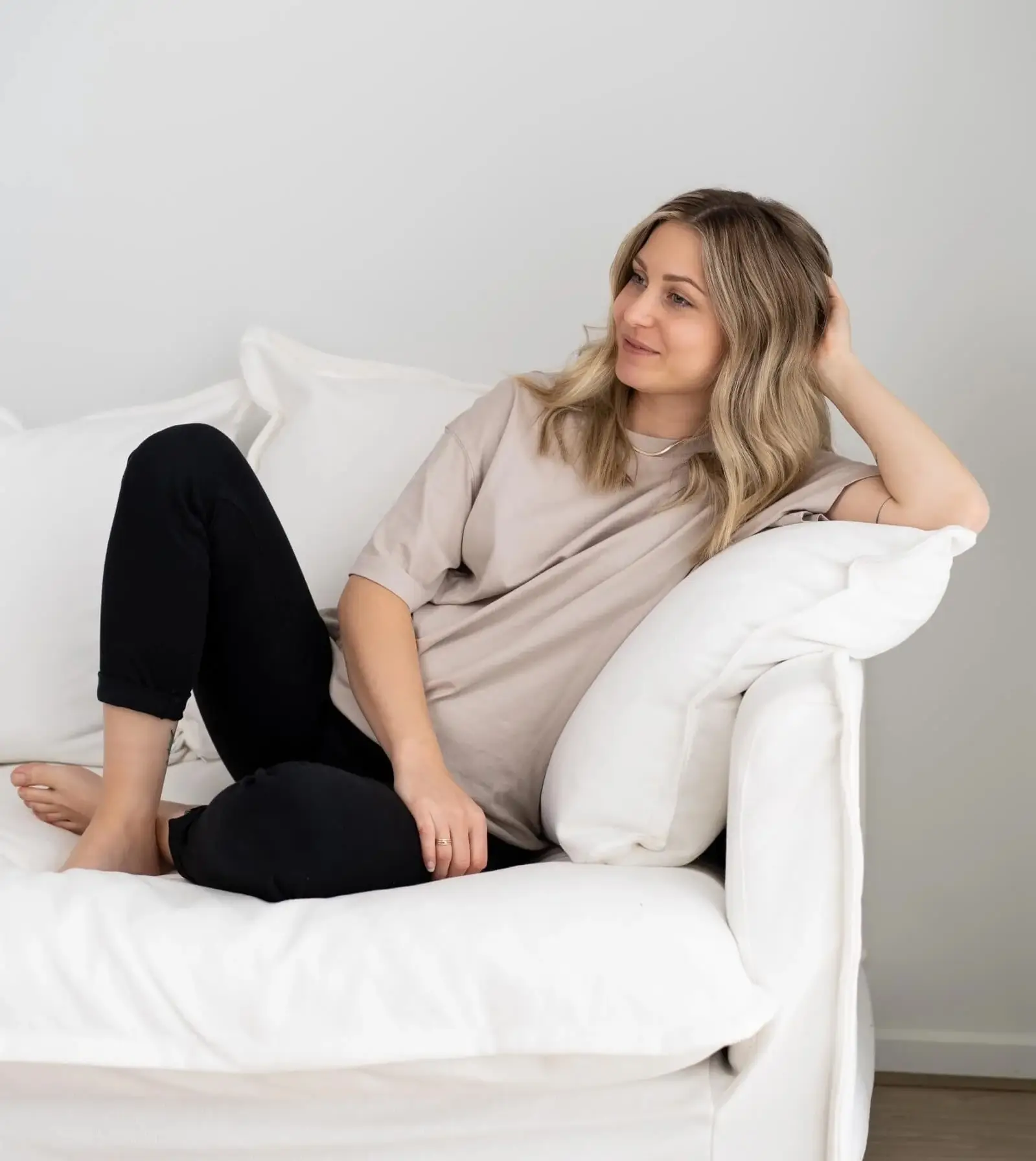 Woman with blonde hair wearing a beige shirt and black pants lounging on a white sofa, resting her head on a white pillow.