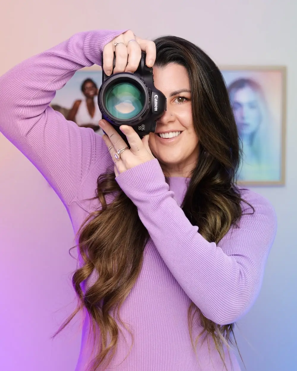 Woman with long brown hair wearing a lavender sweater smiles while holding a Canon camera up to her face.