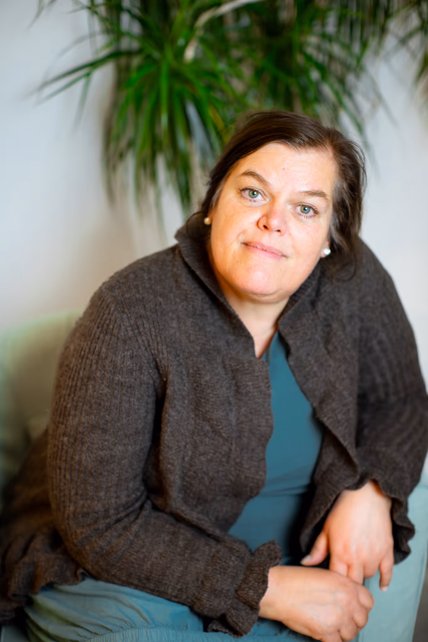 Trude med mørkt hår og grønnblå øyne sitter i en grå lykkegenser foran en stor grønn plante.