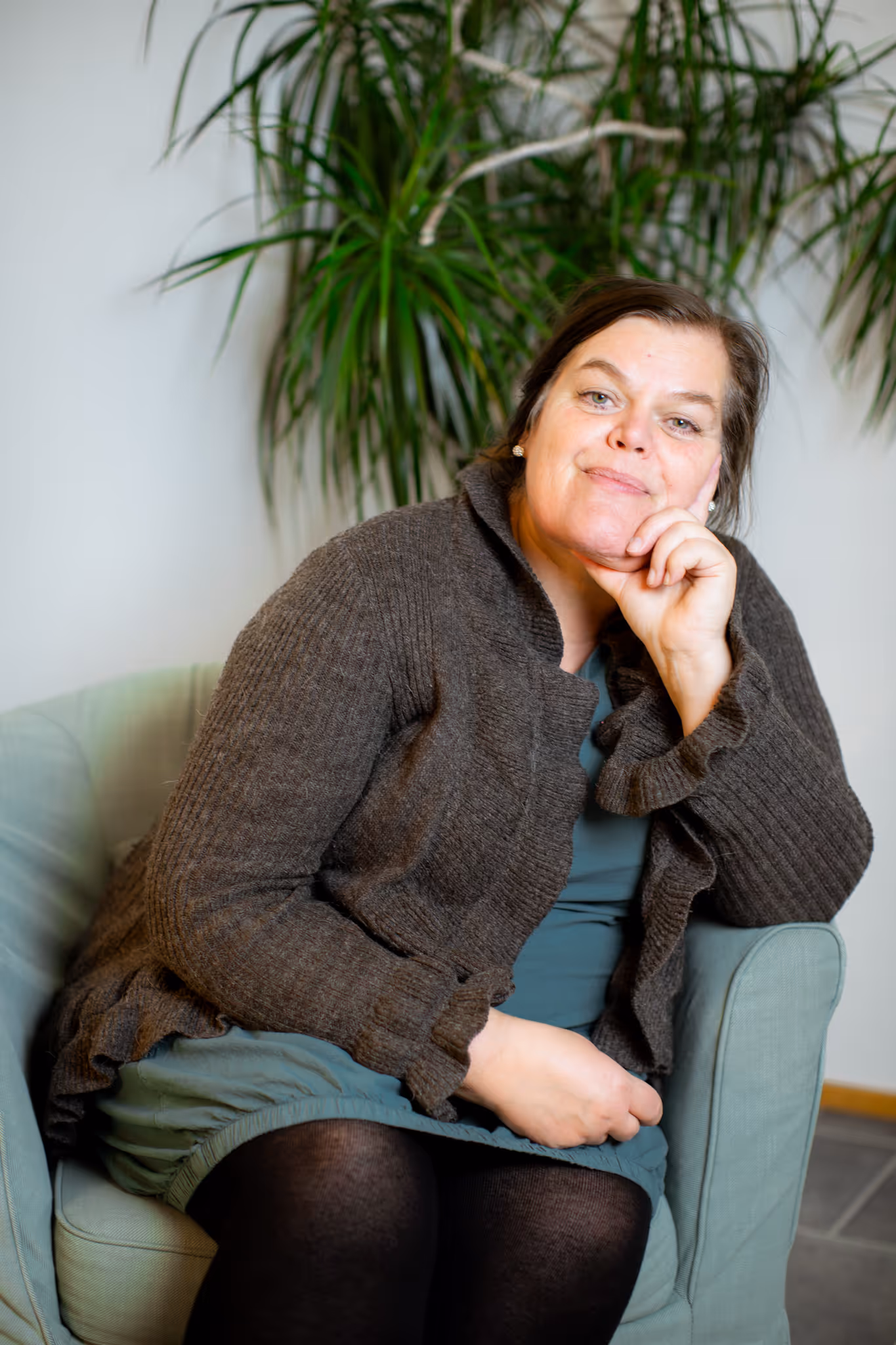 Trude i brun ulljakke sittende avslappet i en grønn stol med en stor grønn plante i bakgrunnen.