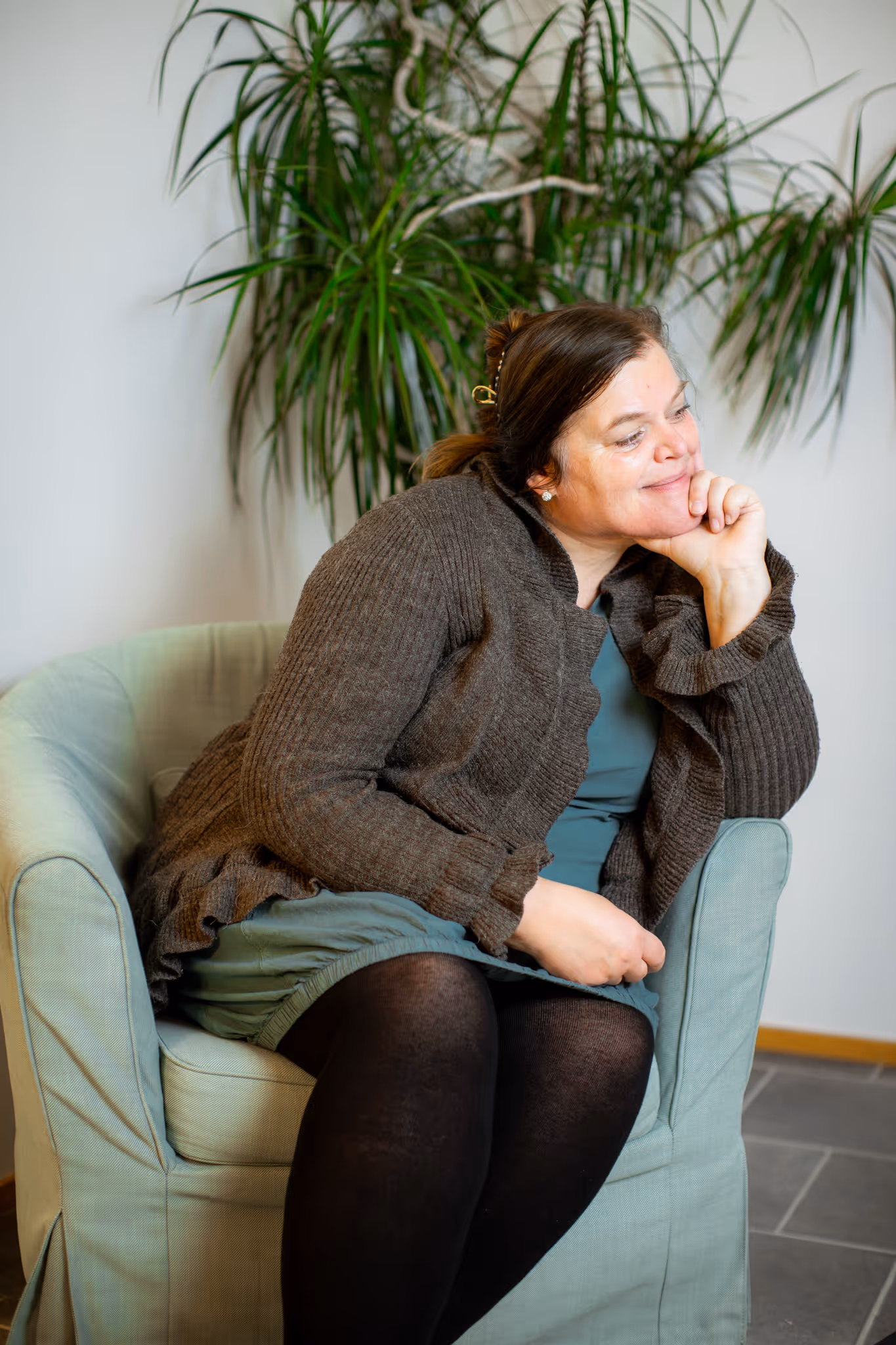Trude som sitter i en lett grønn lenestol, iført en brun strikket genser og svart strømpebukse, smilende med hodet hvilende på hånden.