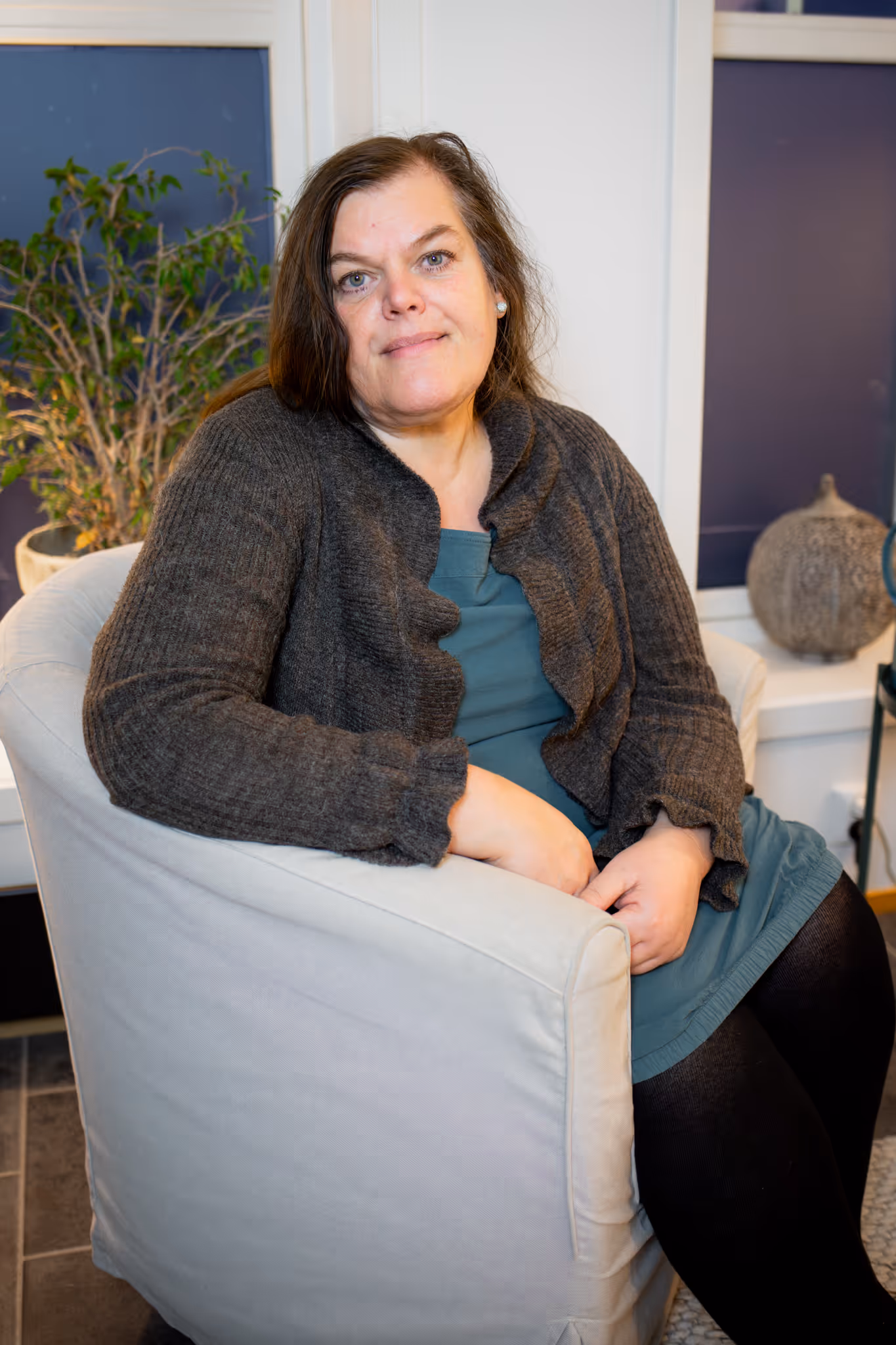 Trude med mørkt, skulderlangt hår sitter i en lys lenestol med en grønn kjole og en mørk grå cardigan.