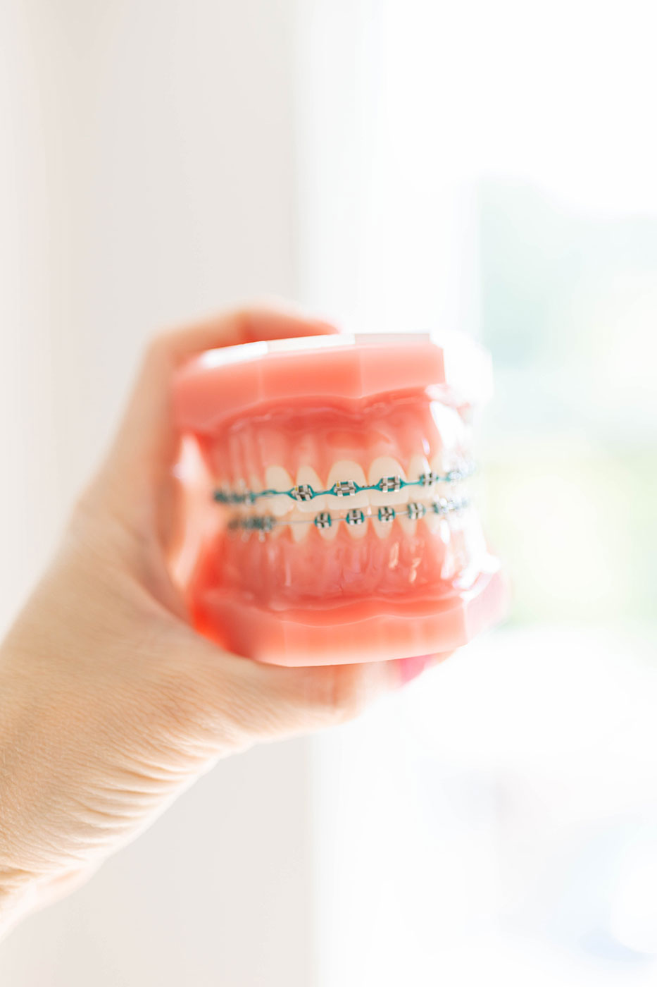 A hand holding a dental model fitted with traditional metal braces and a teal archwire