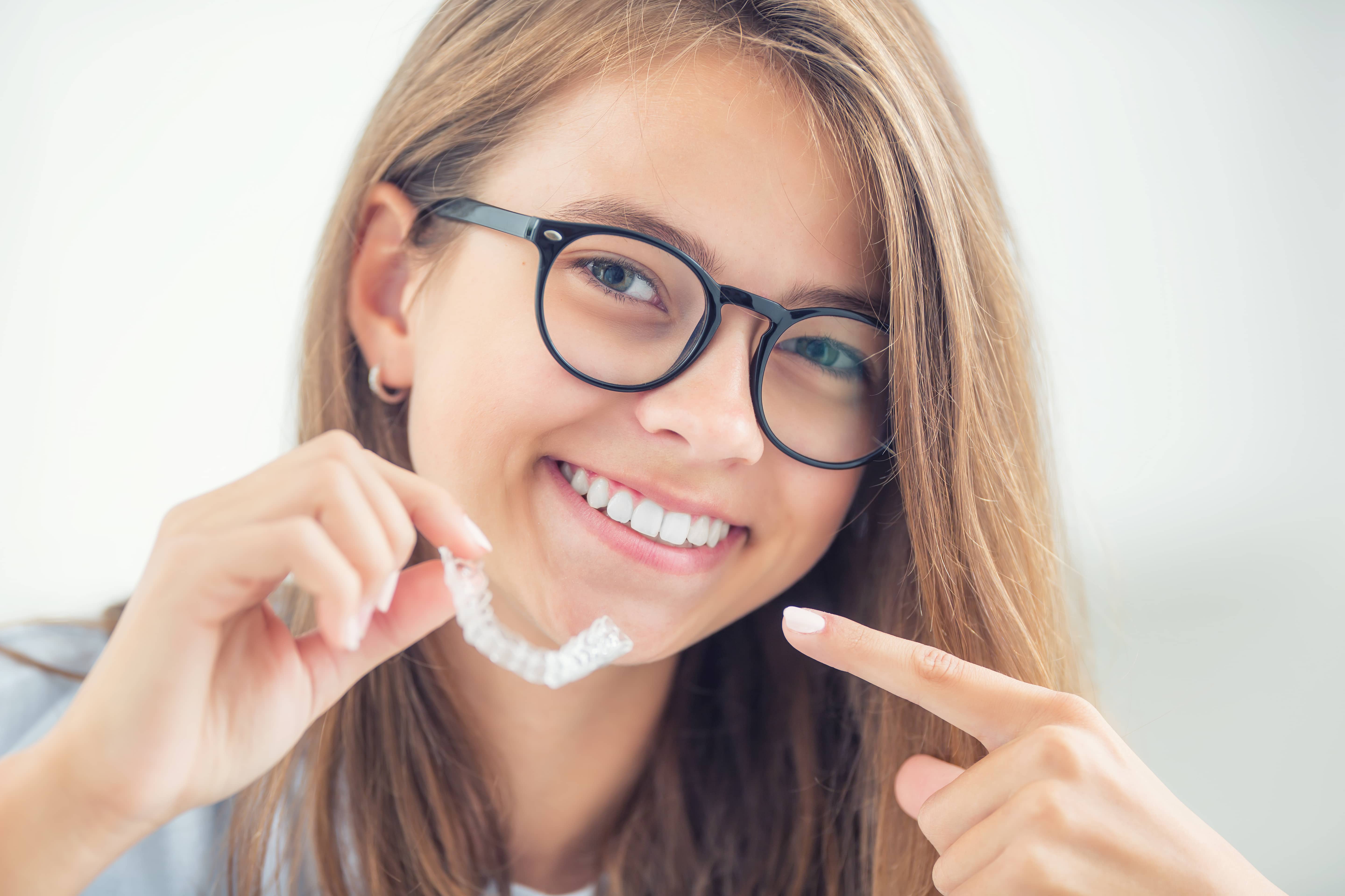 A smiling teenage girl holding a clear Invisalign aligner and pointing at her straight teeth