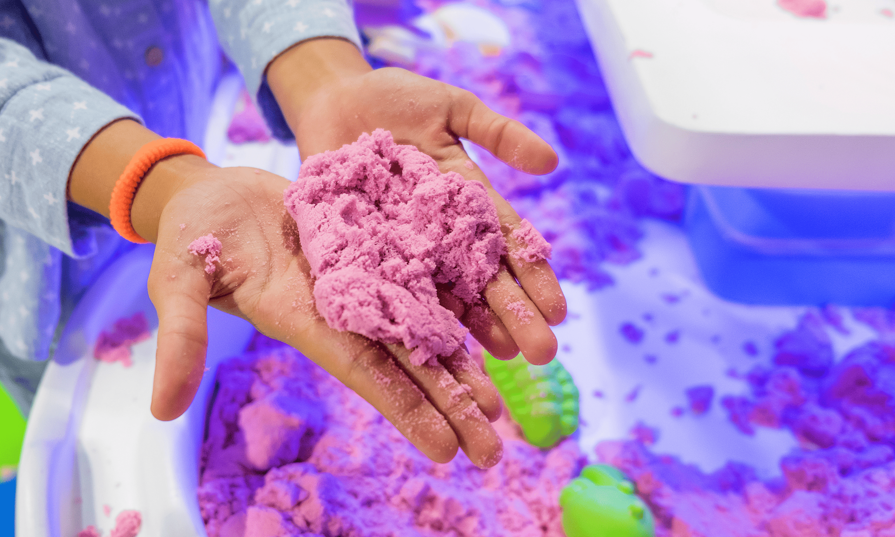 Child holding pink kinetic sand in their hands over a play area filled with more pink sand and green sand molds.