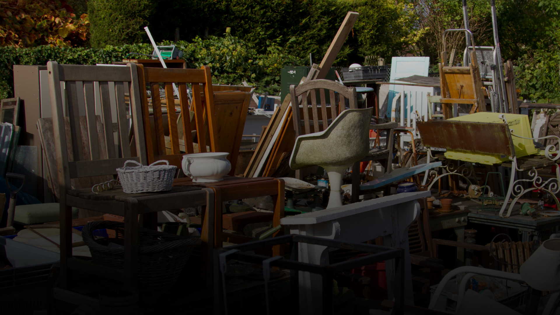 Outdoor scene with a cluttered assortment of old wooden chairs, a white swivel chair, baskets, and various other vintage furniture and items.
