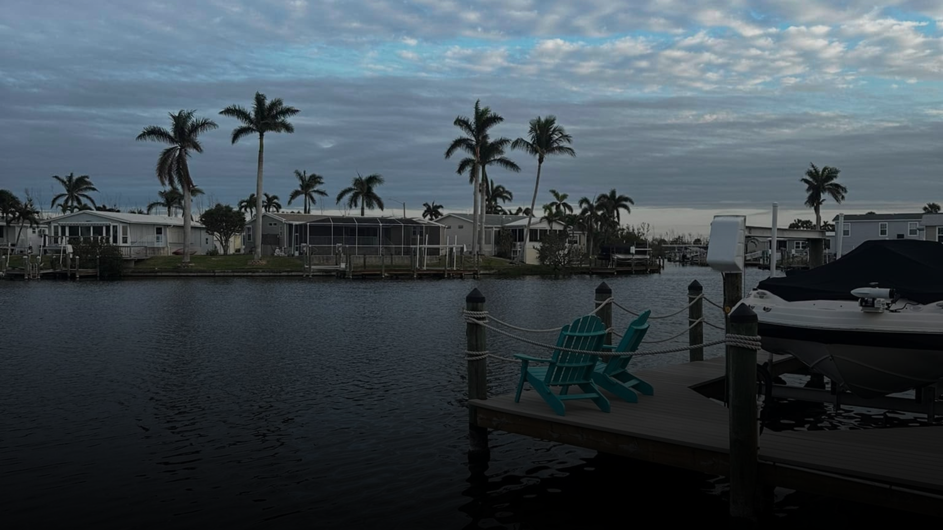 Waterfront scene with palm trees, houses in the background, two teal chairs on a dock, and a covered boat moored alongside.