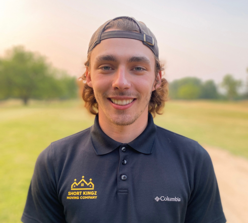 Young man wearing a backward cap and navy polo shirt with Short Kingz Moving Company and Columbia logos standing outdoors on a grassy area.
