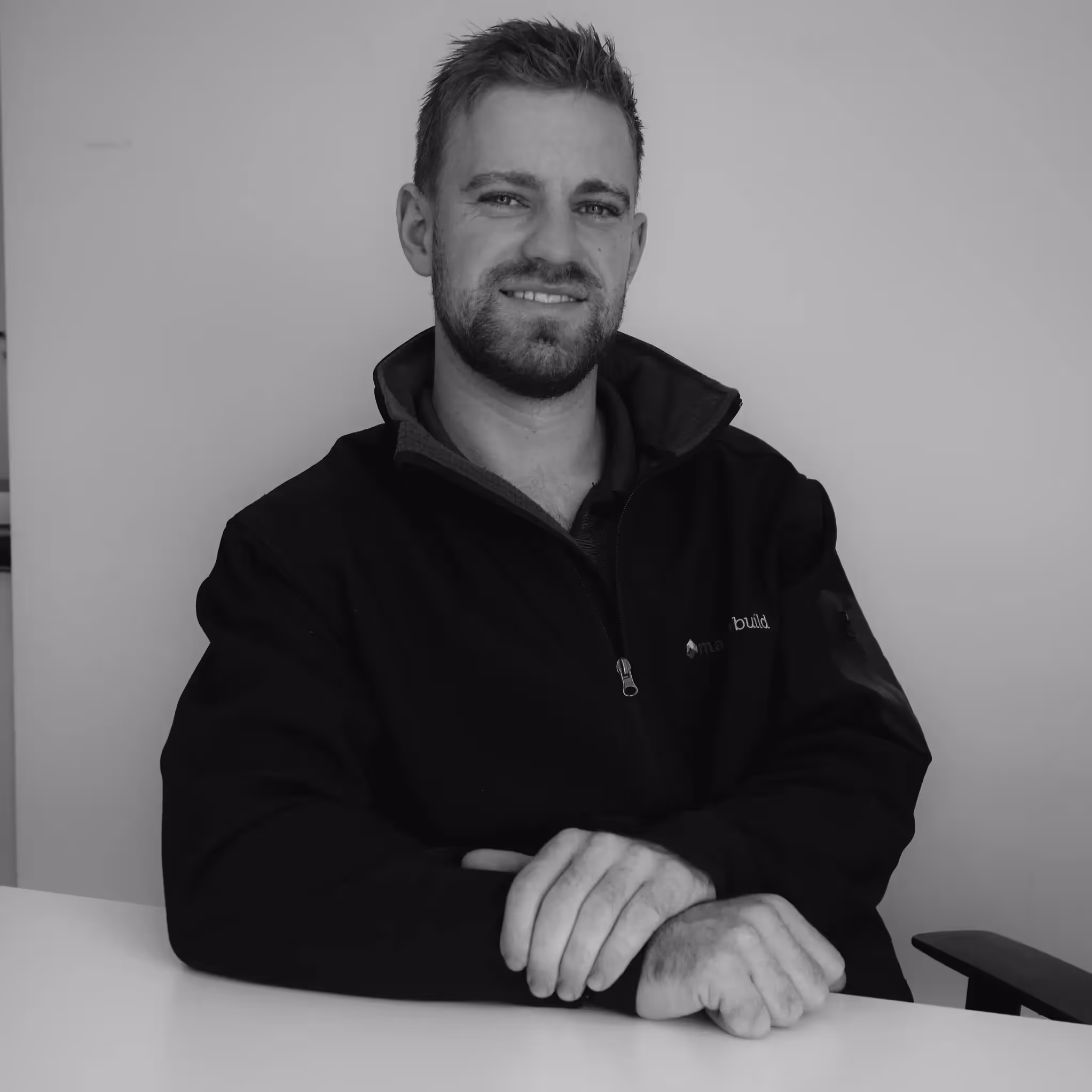 A black and white photo of a man with short hair. His arm rests on a table