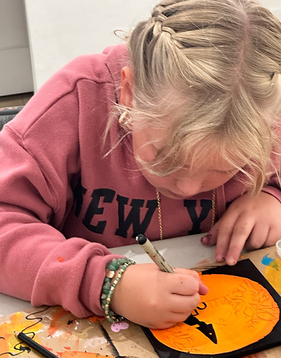 Young girl with braided blonde hair coloring an orange paper pumpkin with a black marker.