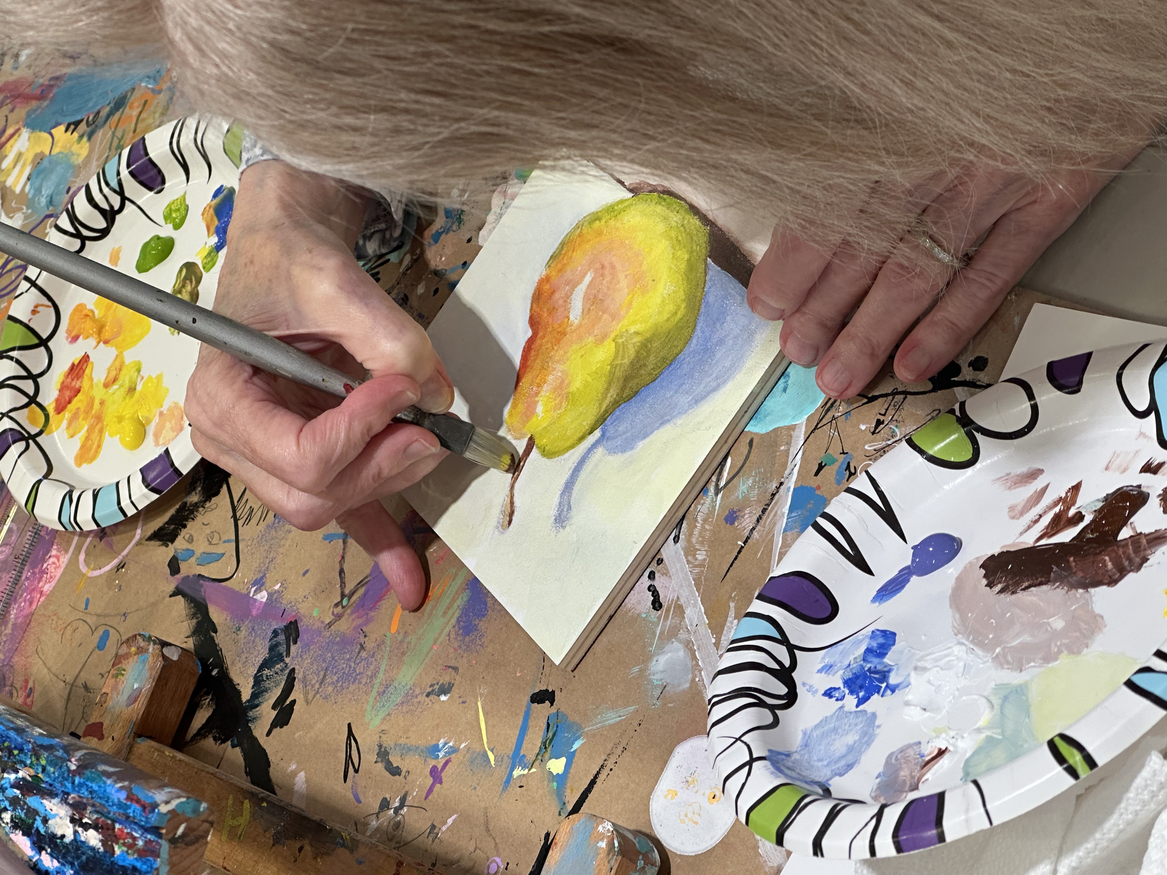 Person painting a yellow and orange pear with a brown stem on white paper using a paintbrush, surrounded by paint palettes and a paint-covered table.