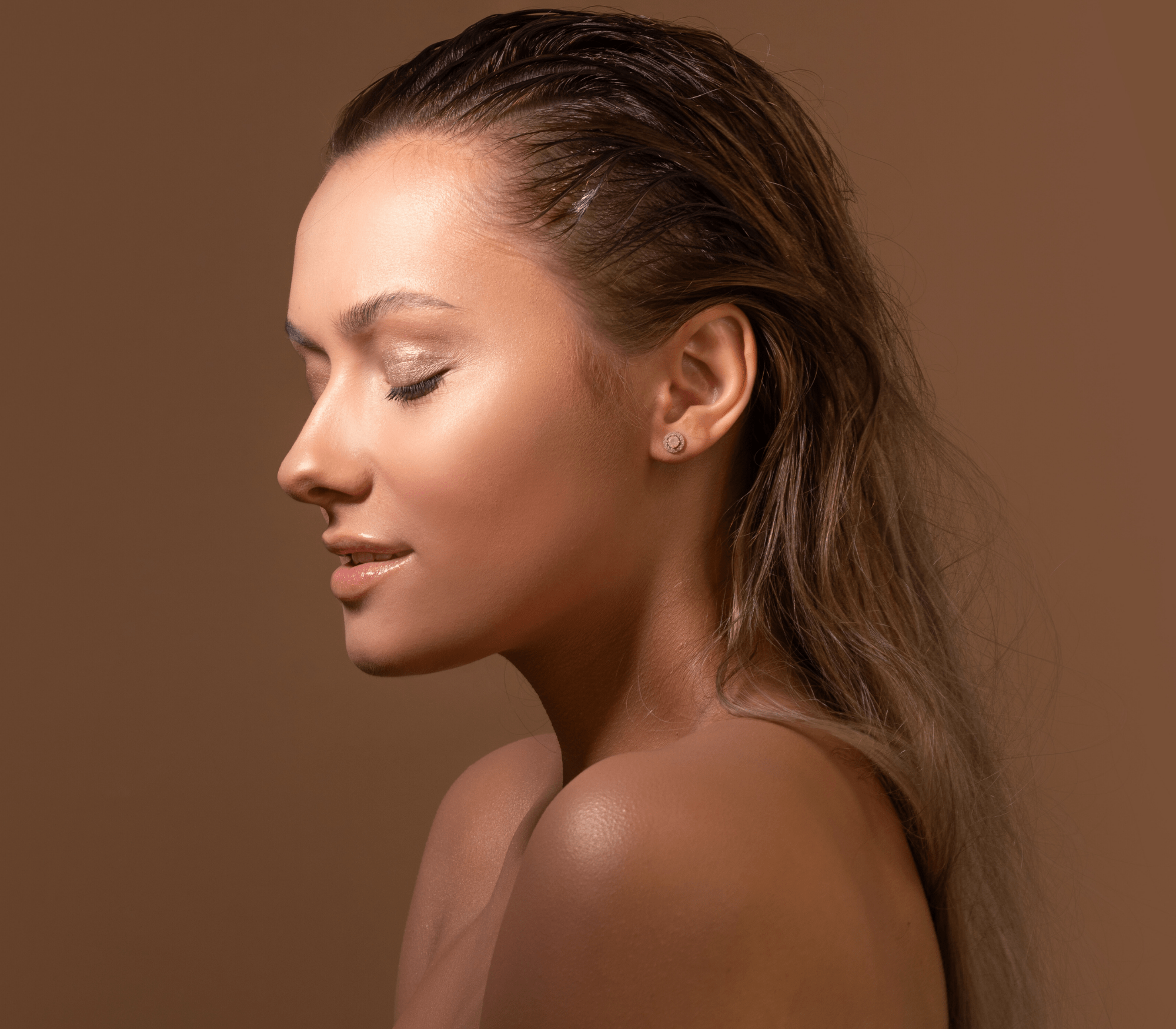 Profile of a woman with sleek wet hair, closed eyes, and glossy lips against a brown background posing for TANNED TO - Automated spray tan service in Toronto.